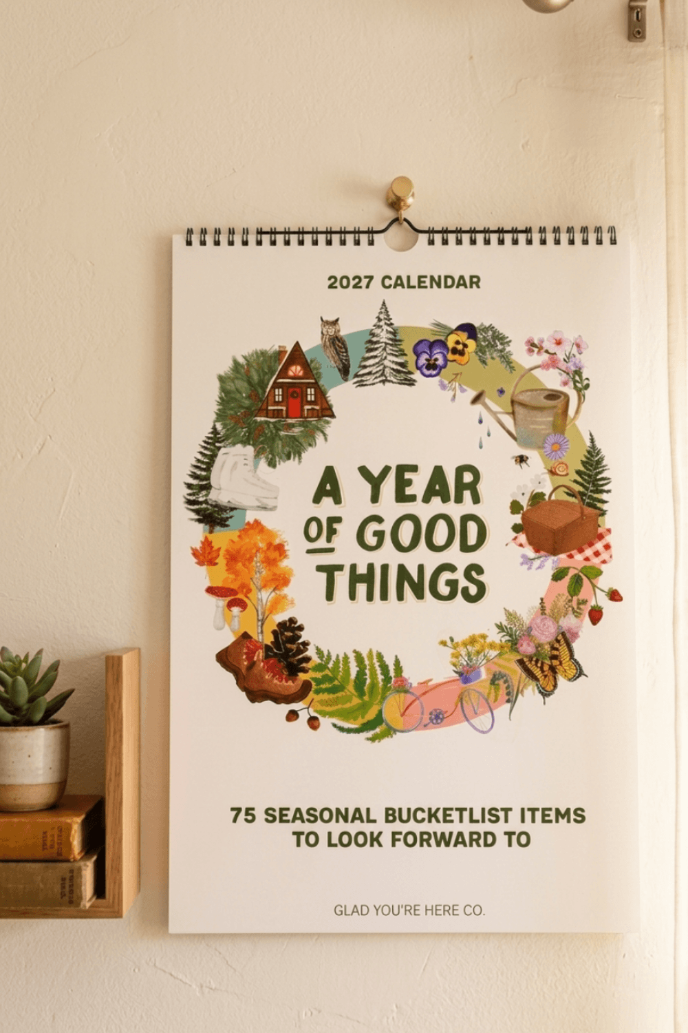 A 2027 spiral-bound wall calendar hanging on a beige wall, featuring a circular illustrated wreath of autumn and nature elements around the text 'A YEAR OF GOOD THINGS' and '75 SEASONAL BUCKETLIST ITEMS TO LOOK FORWARD TO.'.