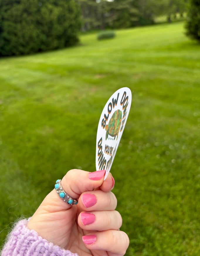 A hand with pink nail polish and turquoise rings holds a white sticker that reads 'SLOW DO NOT OVERLOOK' against a blurred green grassy background.