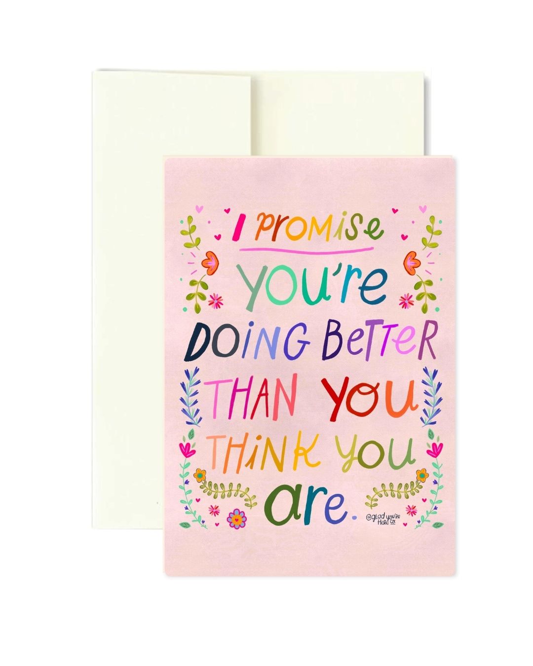A pink greeting card with colorful hand-lettered text reading 'I promise you're doing better than you think you are' surrounded by small floral illustrations, shown with a white envelope behind it on a white background.