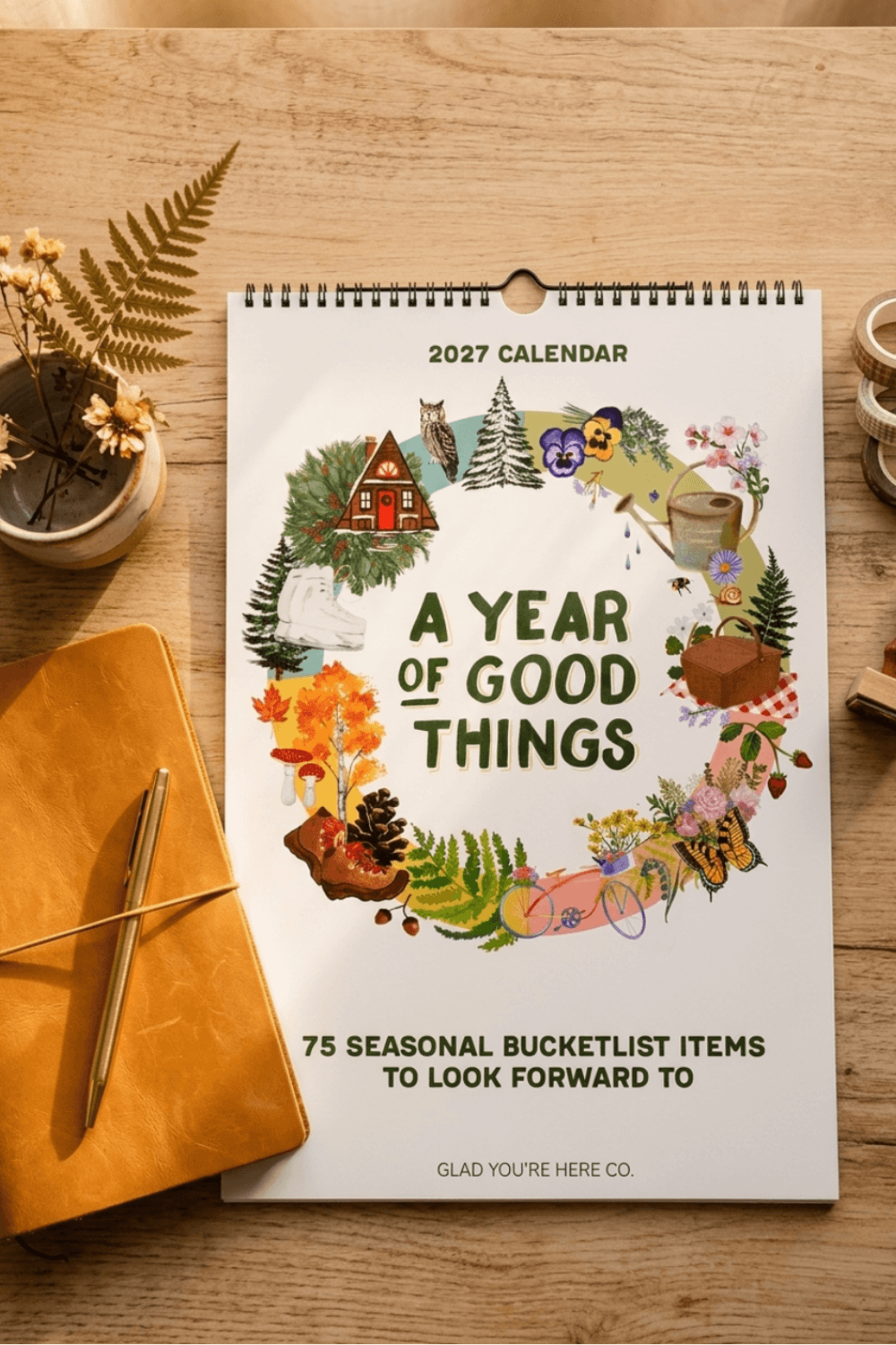 A 2027 spiral-bound calendar titled 'A Year of Good Things' lies on a wooden tabletop, surrounded by a mustard yellow notebook, a pen, dried ferns, and small decorative objects. The calendar cover features a colorful illustrated wreath with seasonal and cozy motifs, including a cabin, owl, pine tree, mug, and flowers.