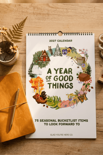 A 2027 spiral-bound calendar titled 'A Year of Good Things' lies on a wooden tabletop, surrounded by a mustard yellow notebook, a pen, dried ferns, and small decorative objects. The calendar cover features a colorful illustrated wreath with seasonal and cozy motifs, including a cabin, owl, pine tree, mug, and flowers.