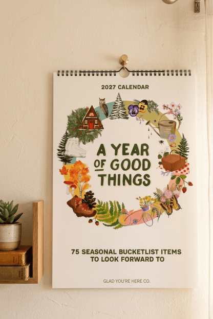 A 2027 spiral-bound wall calendar hanging on a beige wall, featuring a circular illustrated wreath of autumn and nature elements around the text 'A YEAR OF GOOD THINGS' and '75 SEASONAL BUCKETLIST ITEMS TO LOOK FORWARD TO.'.