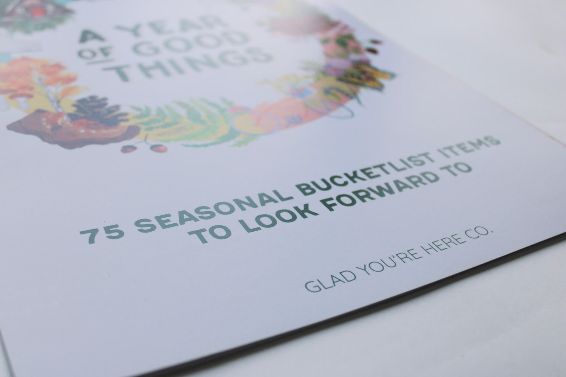 A close-up of a printed card or booklet page with the headline 'A Year of Good Things' and smaller text '75 Seasonal Bucketlist Items to Look Forward To,' plus the brand name 'Glad You're Here Co.' on a white background.
