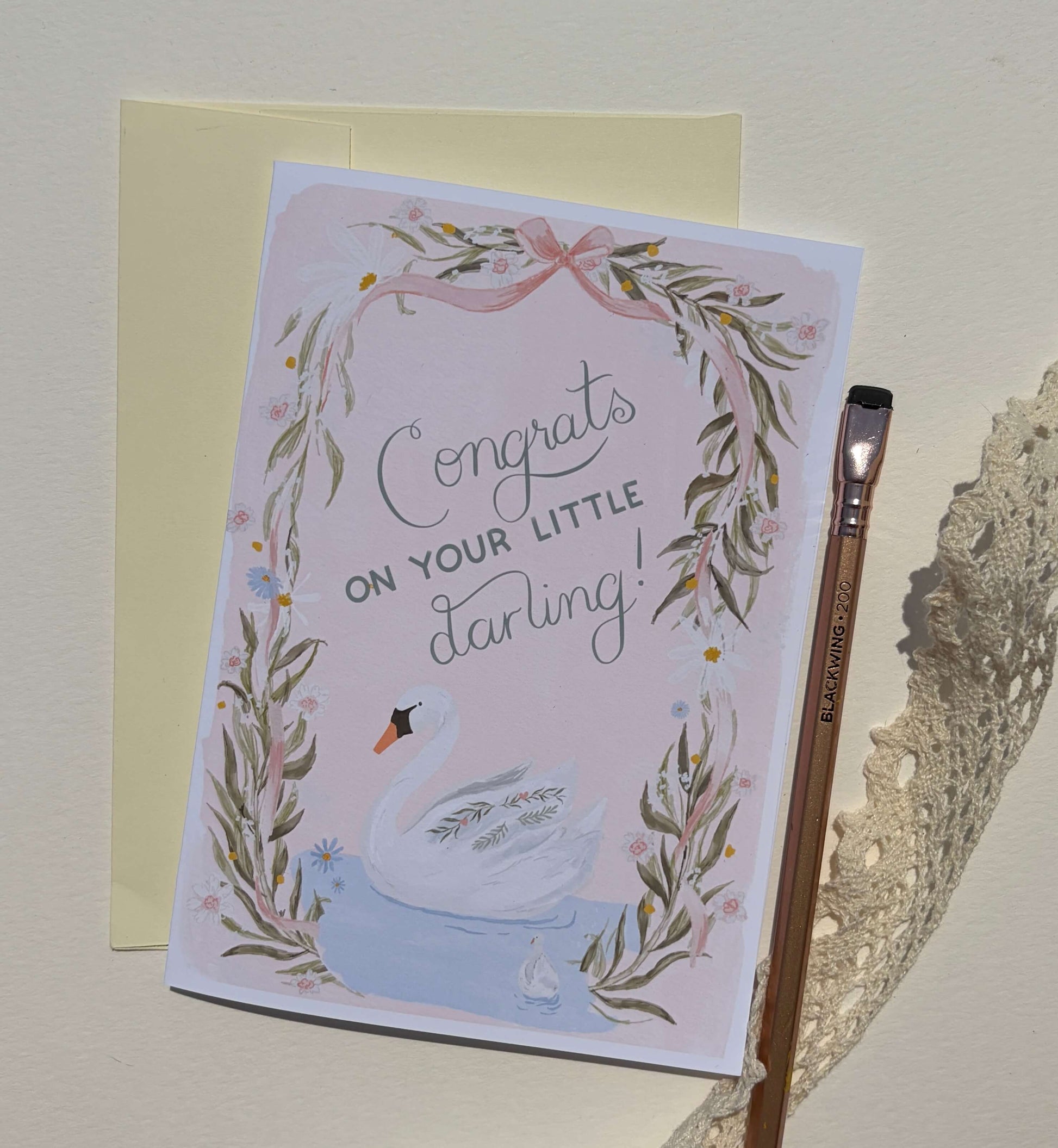 A congratulations card for a baby swan, featuring a watercolor illustration of a white swan in a leafy wreath with the text 'Congrats ON YOUR LITTLE darling!'. The card is shown with a pale yellow envelope behind it and a wooden paintbrush on the right, all set against a light beige textured background.