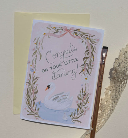 A congratulations card for a baby swan, featuring a watercolor illustration of a white swan in a leafy wreath with the text 'Congrats ON YOUR LITTLE darling!'. The card is shown with a pale yellow envelope behind it and a wooden paintbrush on the right, all set against a light beige textured background.