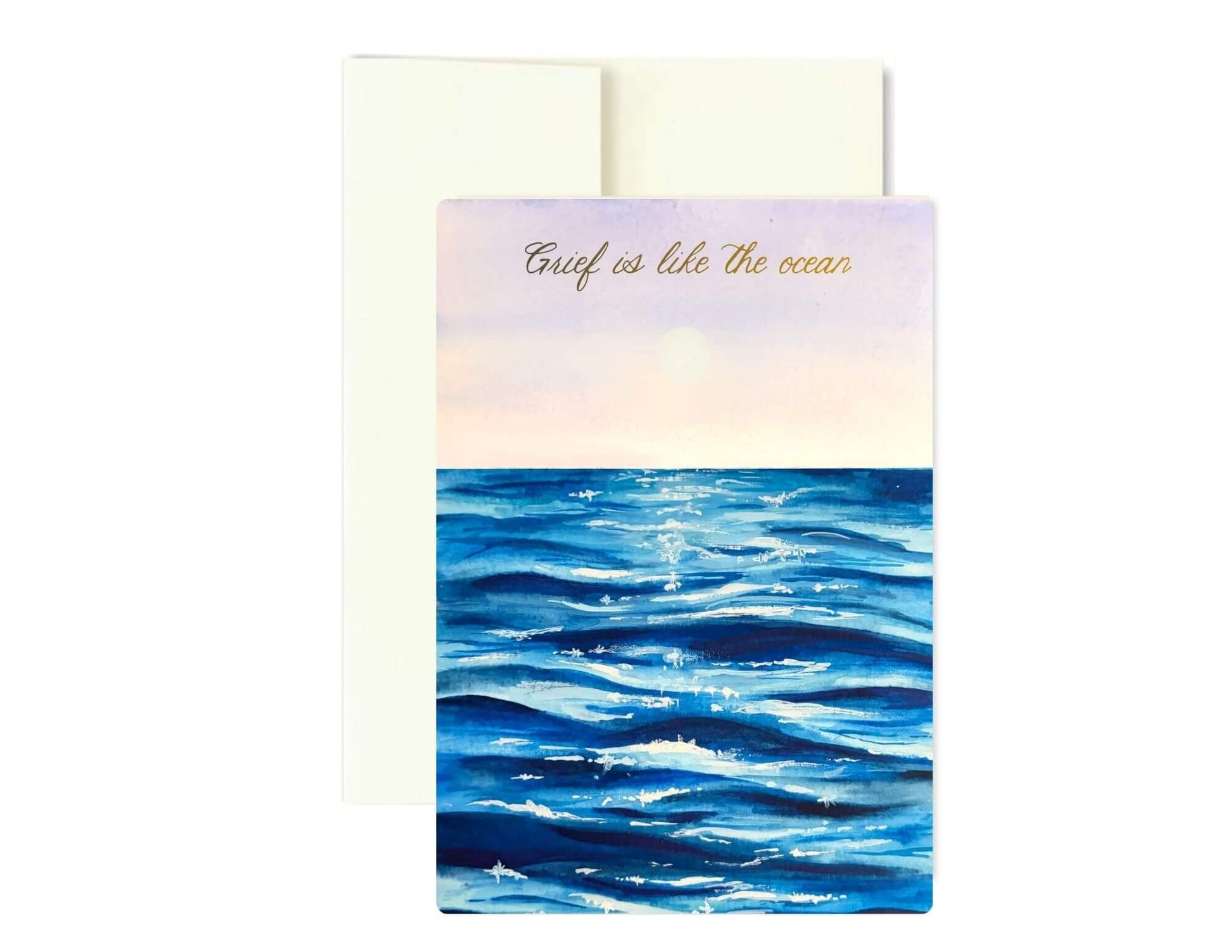 A greeting card with an ocean scene illustration and the words 'Grief is like the ocean' in gold script, shown with a cream envelope behind it on a white background.