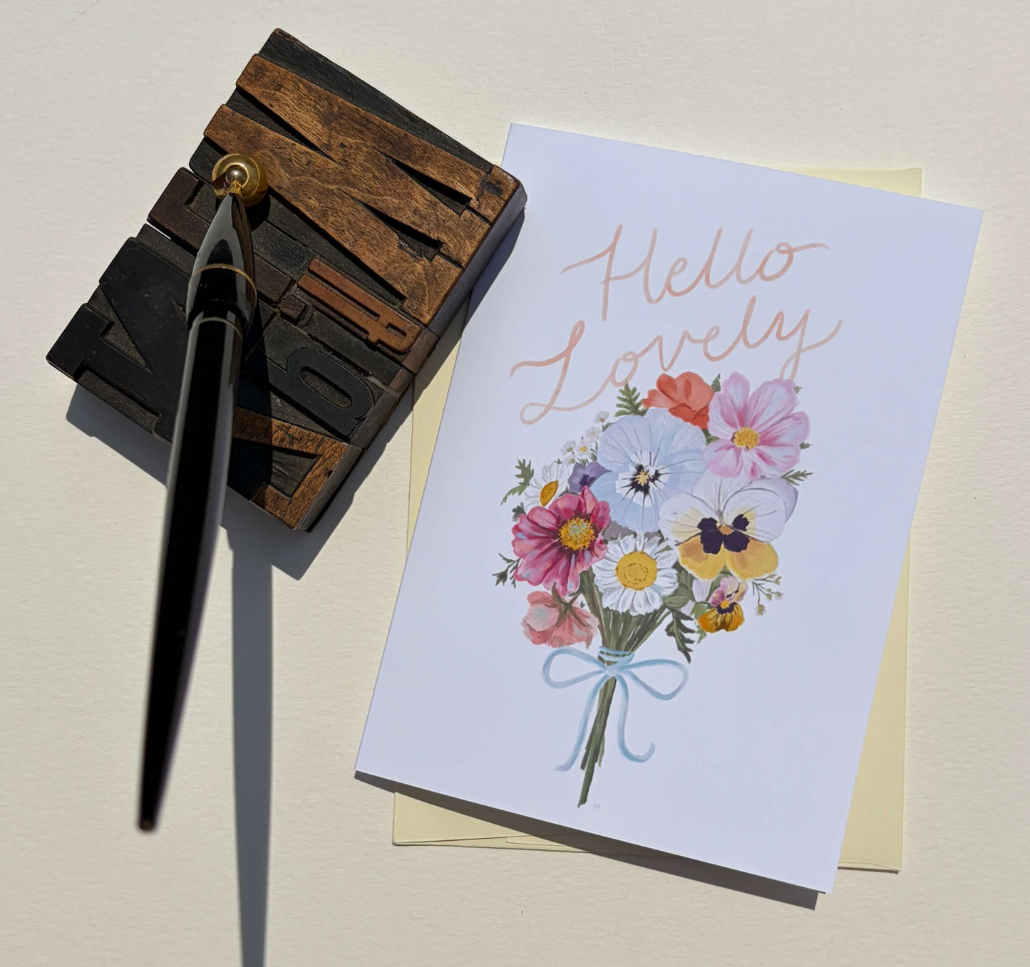 A white greeting card with the words 'Hello Lovely' in rose-gold script and a watercolor bouquet of pink, purple, yellow, and white flowers tied with a light blue ribbon, styled on a cream surface next to a wooden letterpress block and a black pen.
