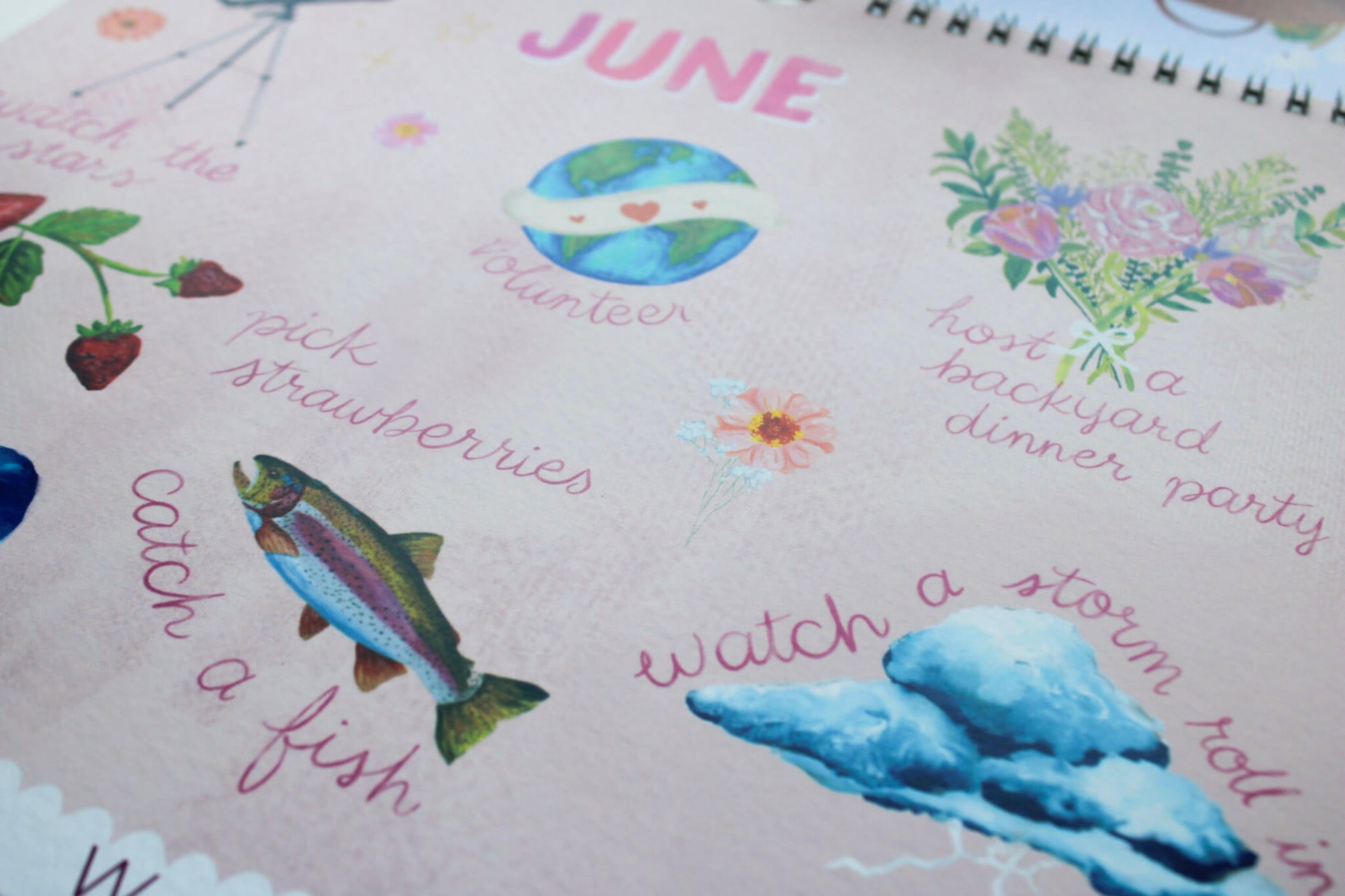 A close-up of a spiral-bound notebook or journal page with a light pink cover, decorated with colorful watercolor illustrations and handwritten text for June, including a fish, strawberries, flowers, and phrases like 'catch a fish' and 'watch a storm roll in'.