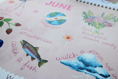 A close-up of a spiral-bound notebook or journal page with a light pink cover, decorated with colorful watercolor illustrations and handwritten text for June, including a fish, strawberries, flowers, and phrases like 'catch a fish' and 'watch a storm roll in'.