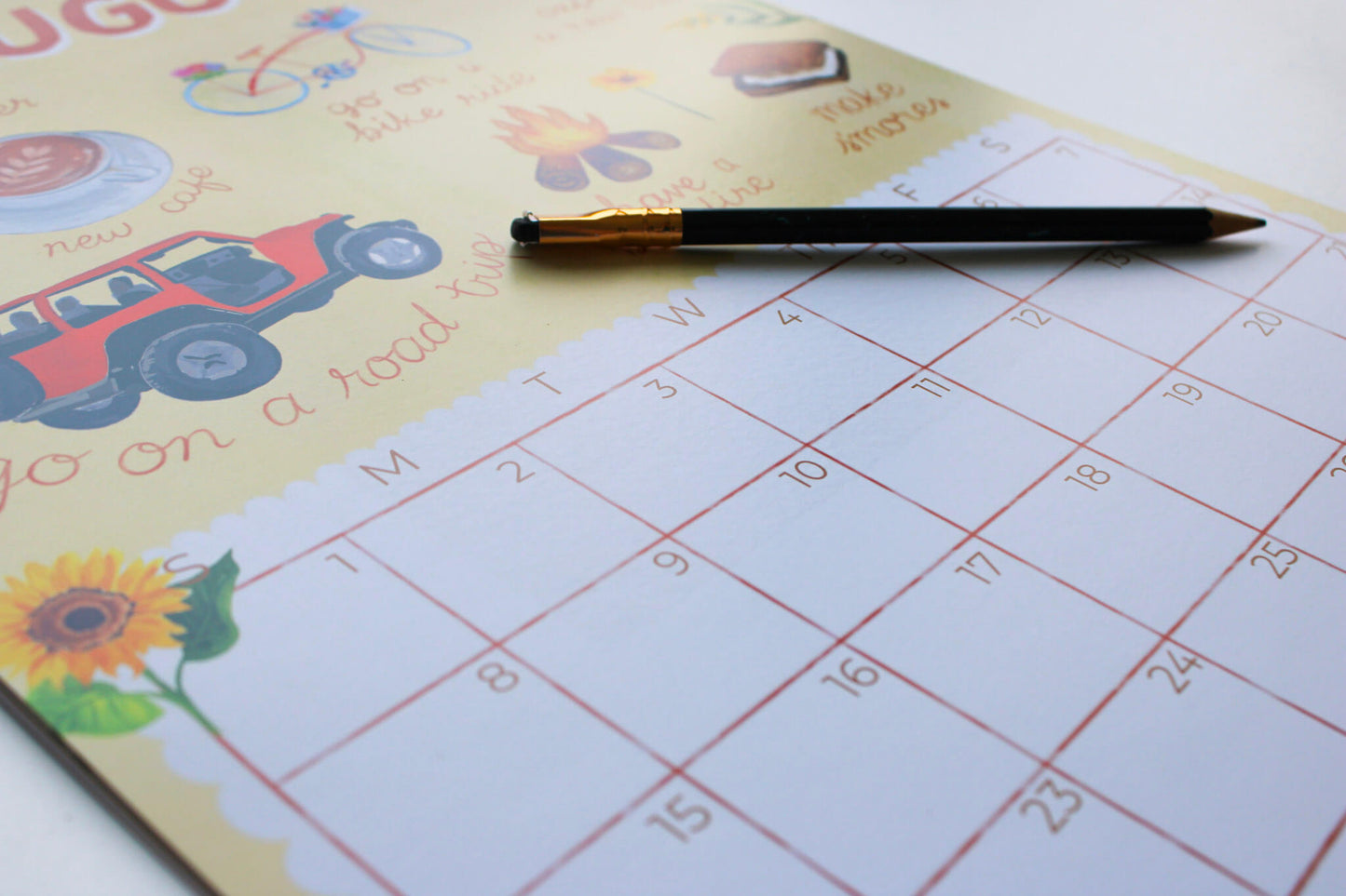 A close-up of a printed monthly calendar pad with a black pencil resting across the top, showing a sunflower and road-trip illustrations on a pale yellow page.