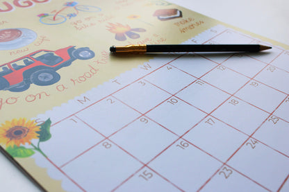 A close-up of a printed monthly calendar pad with a black pencil resting across the top, showing a sunflower and road-trip illustrations on a pale yellow page.