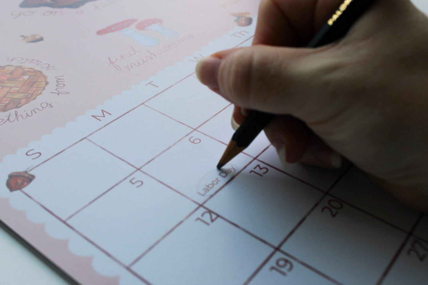 A close-up of a hand holding a black pen and writing in a pink monthly calendar planner with cute illustrations.