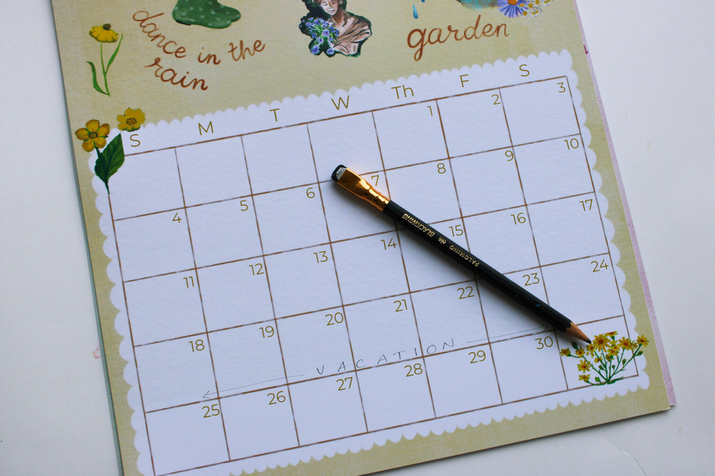 A weekly planner calendar page titled 'dance in the rain garden' with a black pencil resting on it, photographed on a white surface.