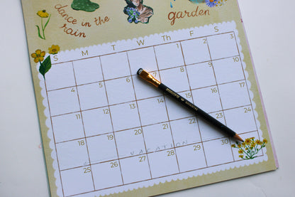 A weekly planner calendar page titled 'dance in the rain garden' with a black pencil resting on it, photographed on a white surface.