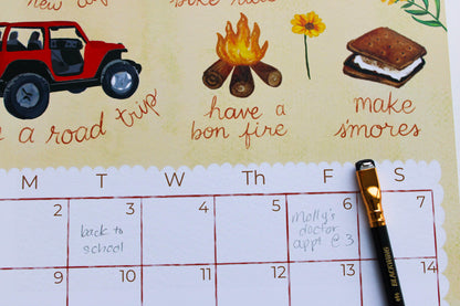 A close-up of a printed weekly planner or calendar page with illustrated icons for road trips, campfires, flowers, and s'mores, plus handwritten notes in the boxes. A black and gold pen rests on the right side of the page.