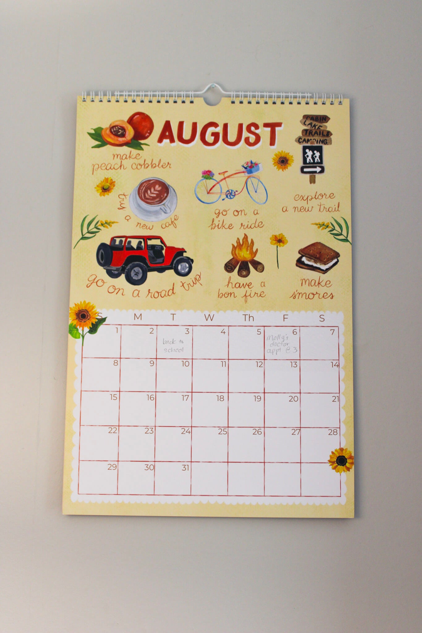 A spiral-bound August wall calendar hanging on a light gray wall. The calendar has a pale yellow background with colorful summer-themed illustrations, including a red jeep, bicycle, campfire, sunflowers, and handwritten notes.