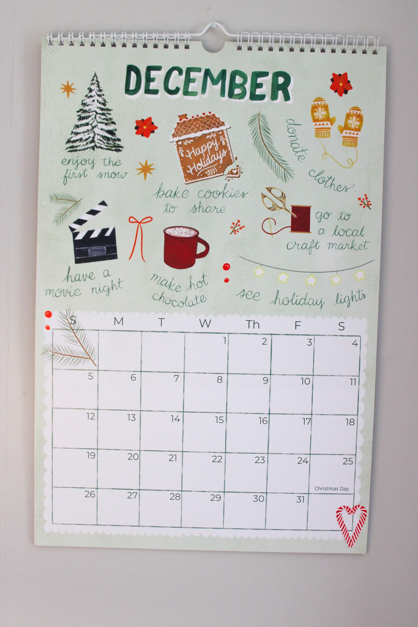 A spiral-bound December calendar page with festive holiday illustrations and handwritten-style holiday phrases, photographed hanging against a light wall.