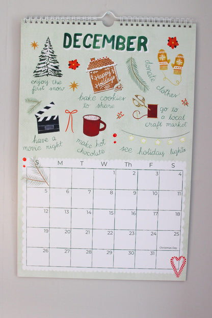 A spiral-bound December calendar page with festive holiday illustrations and handwritten-style holiday phrases, photographed hanging against a light wall.