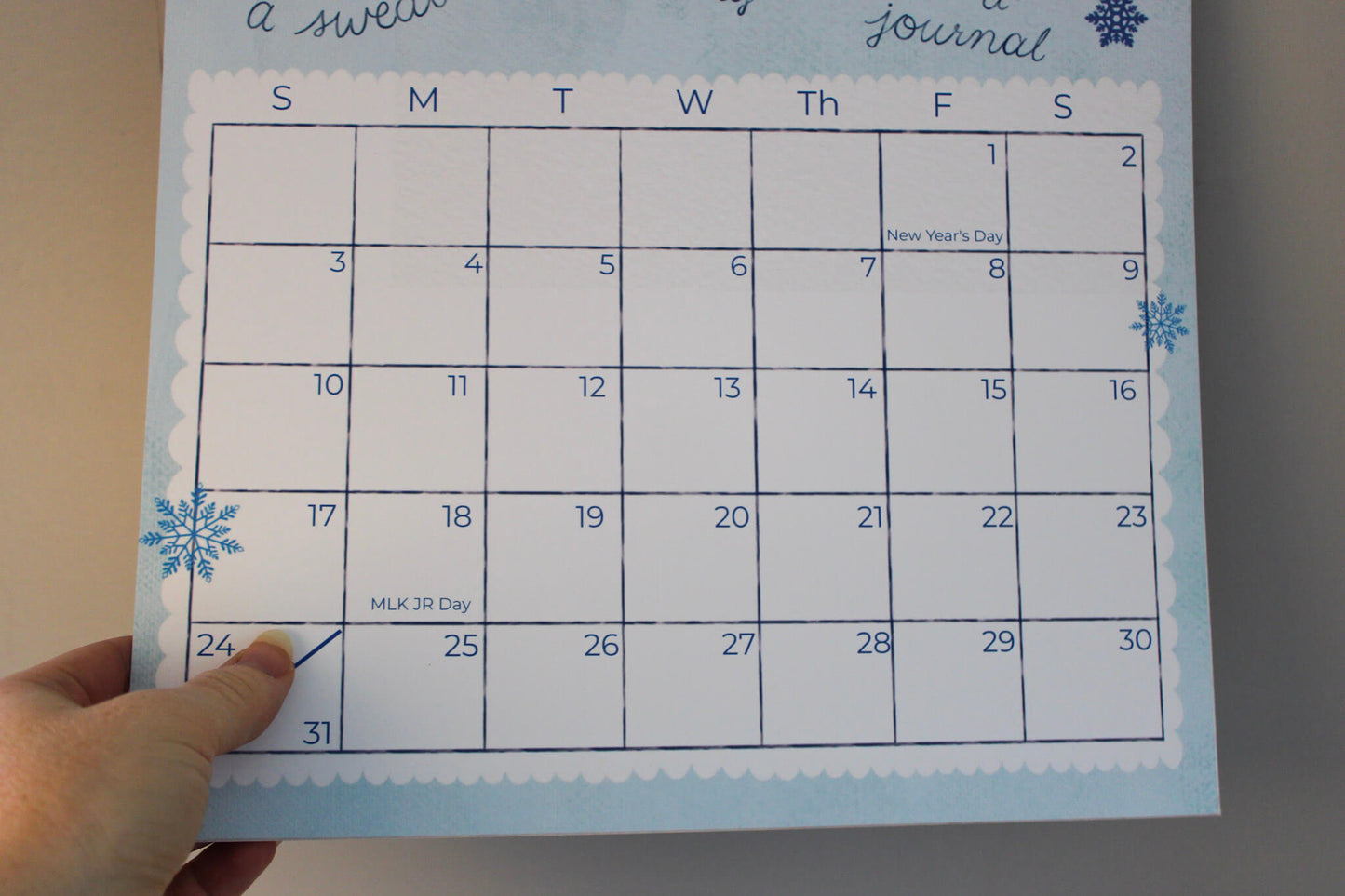 A hand is holding a January 2025 weekly journal calendar page with a winter theme, featuring blue snowflakes and a white scalloped border on a light blue background.