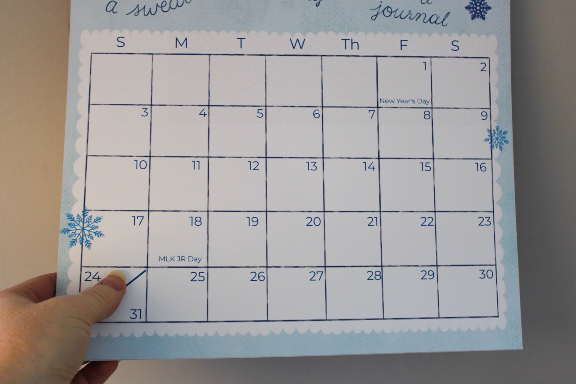 A hand is holding a January 2025 weekly journal calendar page with a winter theme, featuring blue snowflakes and a white scalloped border on a light blue background.