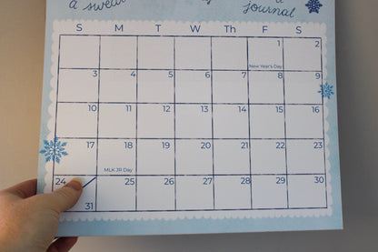 A hand is holding a January 2025 weekly journal calendar page with a winter theme, featuring blue snowflakes and a white scalloped border on a light blue background.