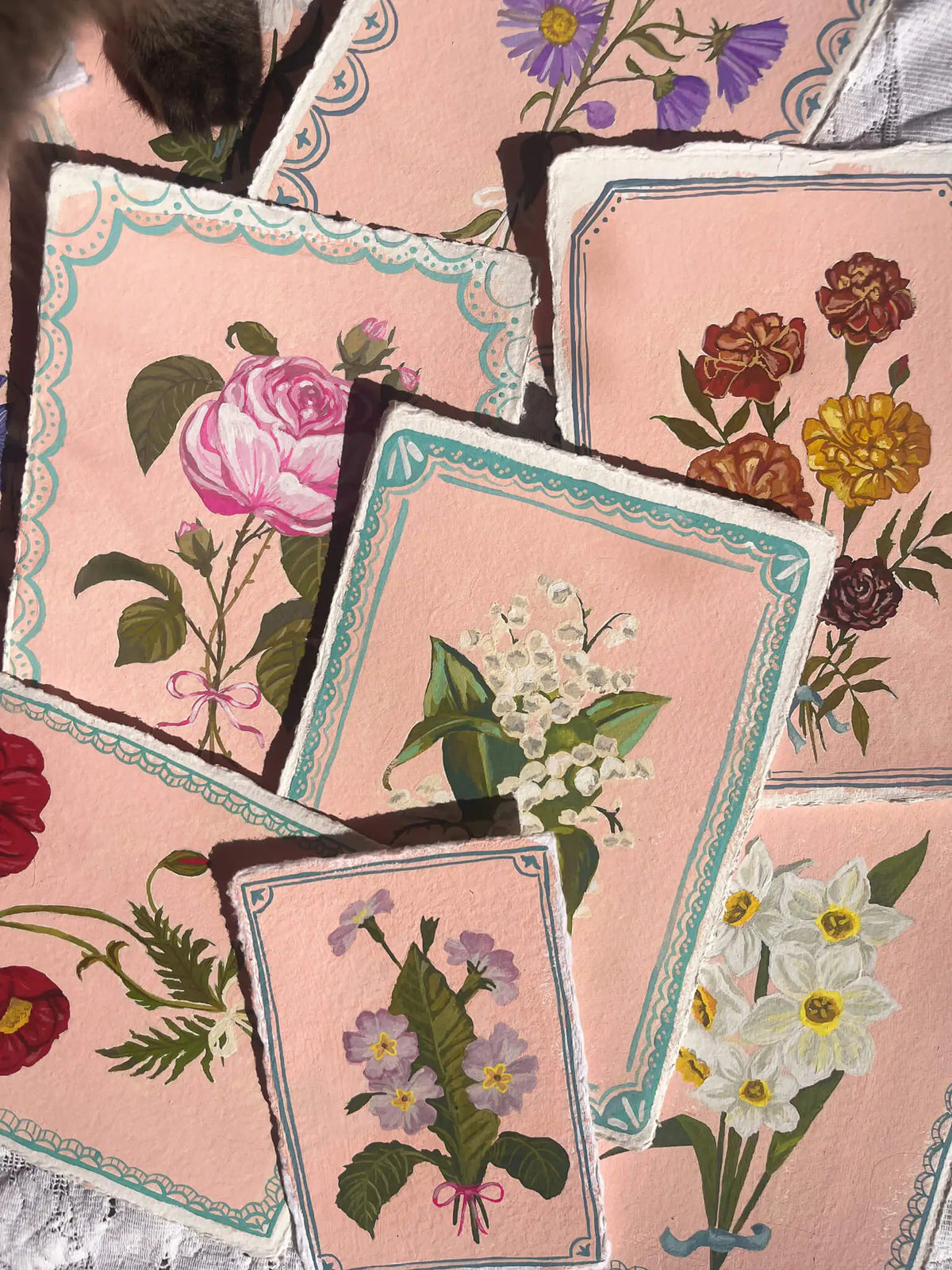 A close-up of several overlapping rectangular cards or prints with pink backgrounds and floral illustrations, including roses, daisies, primroses, and other flowers, arranged on a lace-like surface.