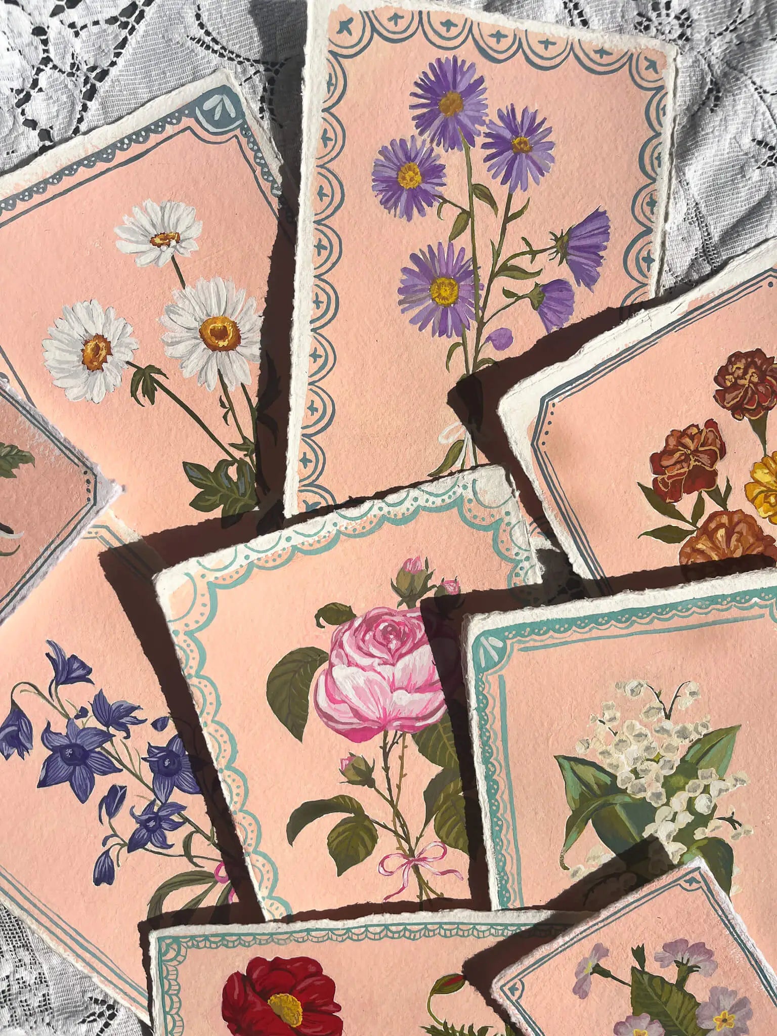 A set of vintage-style floral cards or art prints fanned out on a lace fabric background. The cards feature illustrations of various flowers including daisies, purple asters, a pink rose, bluebells, and lily of the valley, all set against a soft peach-pink background with decorative scalloped borders.