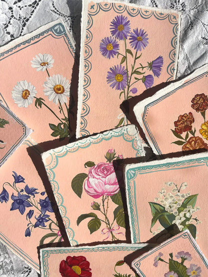 A set of vintage-style floral cards or art prints fanned out on a lace fabric background. The cards feature illustrations of various flowers including daisies, purple asters, a pink rose, bluebells, and lily of the valley, all set against a soft peach-pink background with decorative scalloped borders.