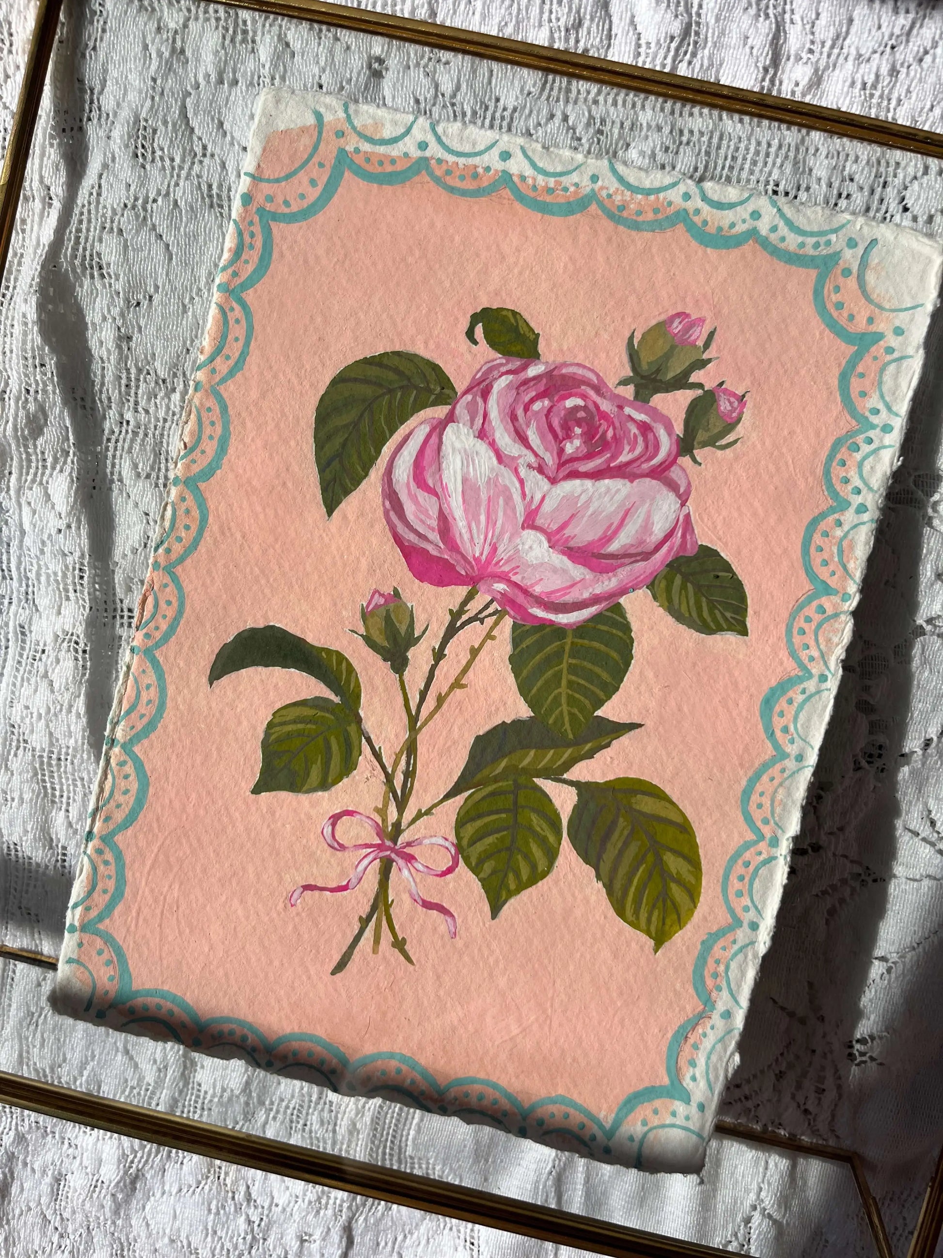 A square decorative art print or card featuring a pink rose with green leaves on a pale pink background, bordered by a scalloped teal and cream pattern. The print is photographed at an angle on a textured gray surface with a gold-toned frame partially visible.