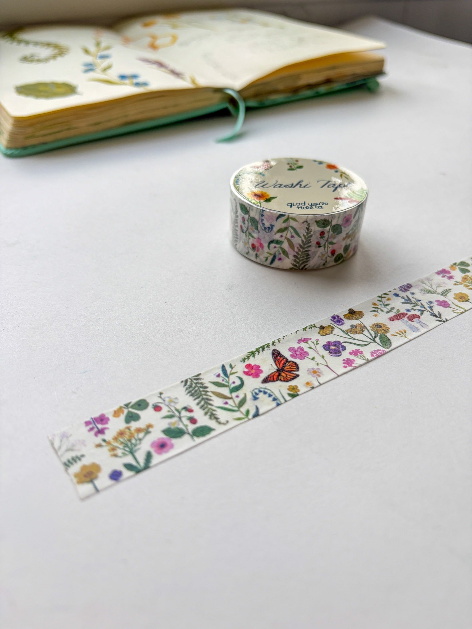Roll of floral washi tape with a strip on a white surface, next to an open notebook.