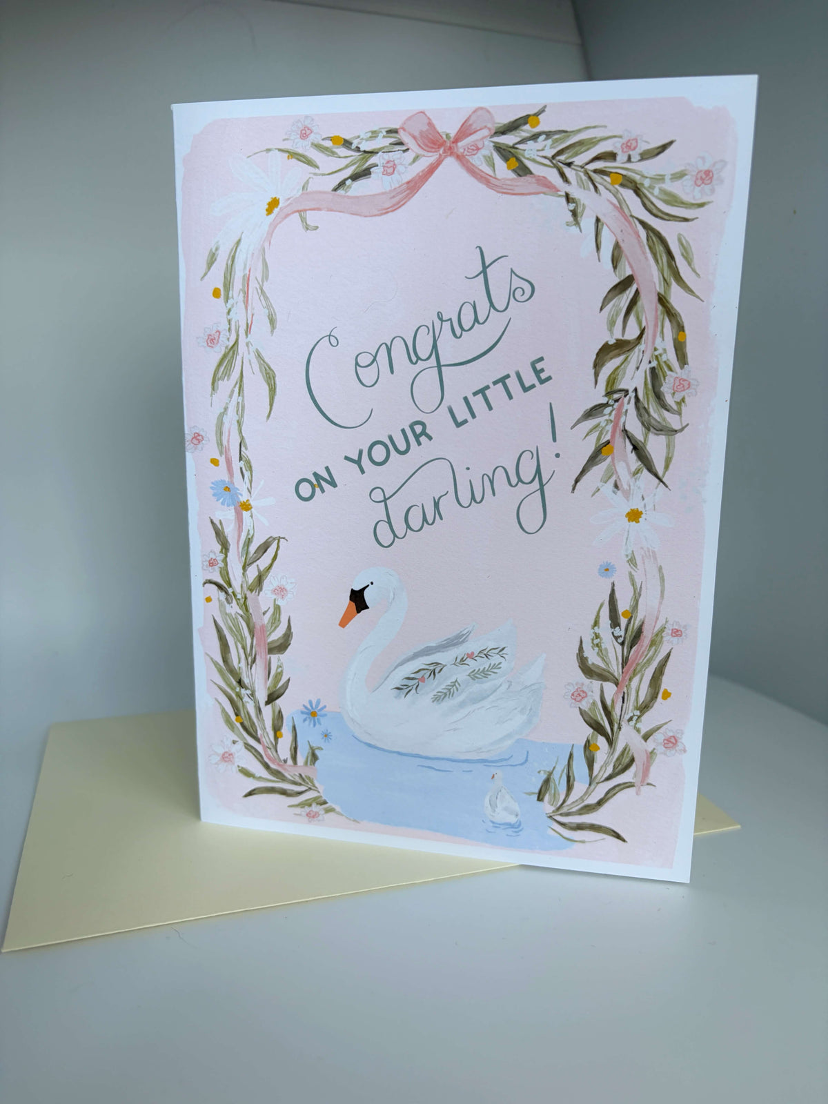 A square greeting card with a white border, featuring a pink background, a swan illustration, and the text 'Congrats on your little darling!'. The card is shown standing upright on a cream envelope against a neutral gray background.