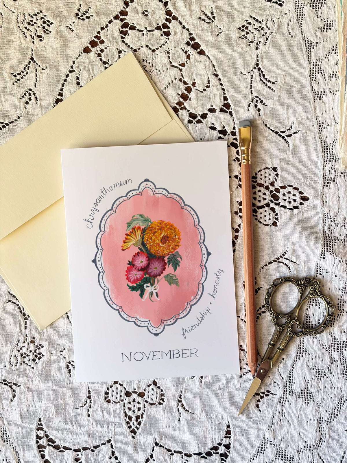 A flat-lay of a botanical monthly card for November, featuring a watercolor illustration of flowers in a pink frame. The card is styled with a pale yellow envelope, a wooden pencil, and ornate scissors on a white lace background.