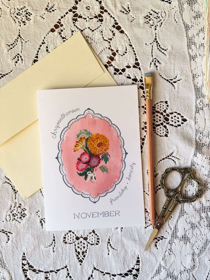 A flat-lay of a botanical monthly card for November, featuring a watercolor illustration of flowers in a pink frame. The card is styled with a pale yellow envelope, a wooden pencil, and ornate scissors on a white lace background.