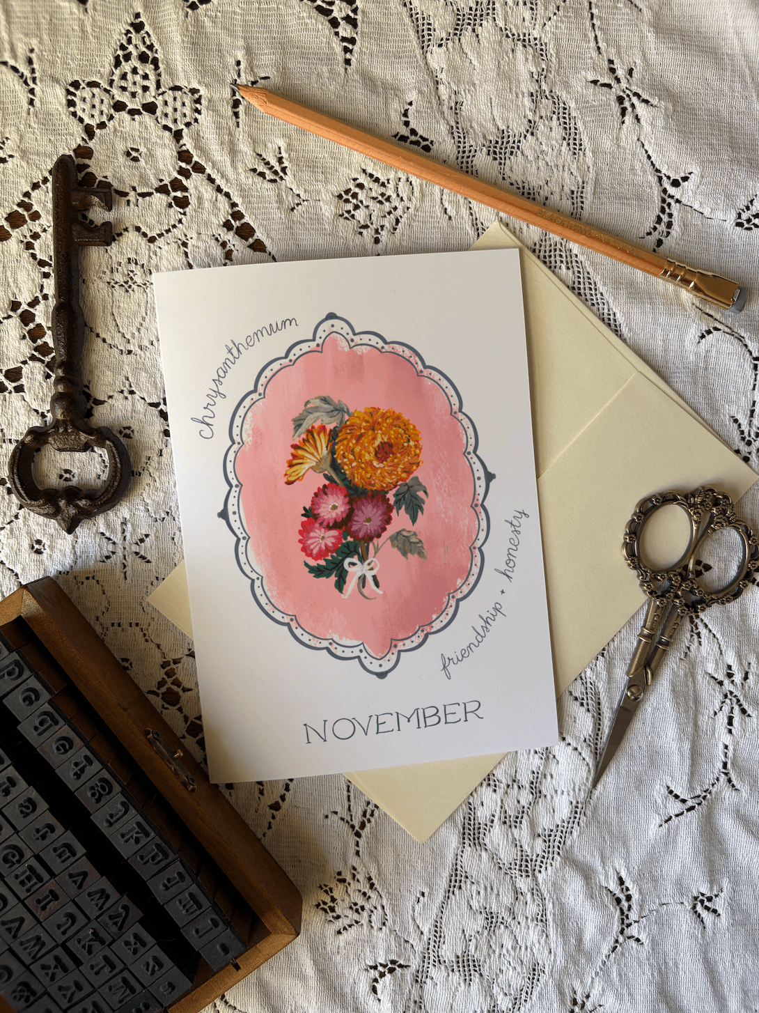 A flat-lay of a handmade monthly greeting card for November, featuring a watercolor floral illustration on a pink background. The card is displayed on a cream envelope, surrounded by vintage-style props including an antique key, a wooden pencil, scissors, and a letterpress box on a white lace fabric background.