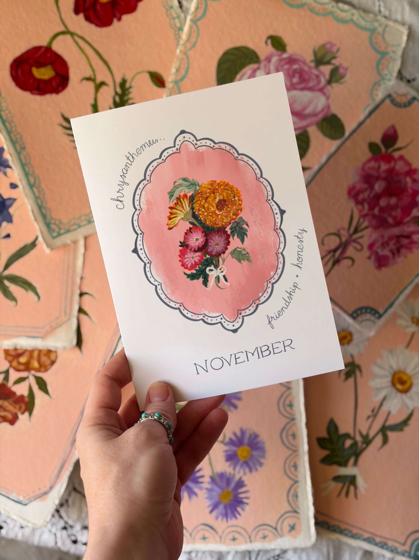 A hand holds a white November card featuring a pink watercolor floral illustration and the words 'Joyworthoman.