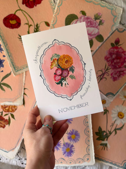 A hand holds a white November card featuring a pink watercolor floral illustration and the words 'Joyworthoman.
