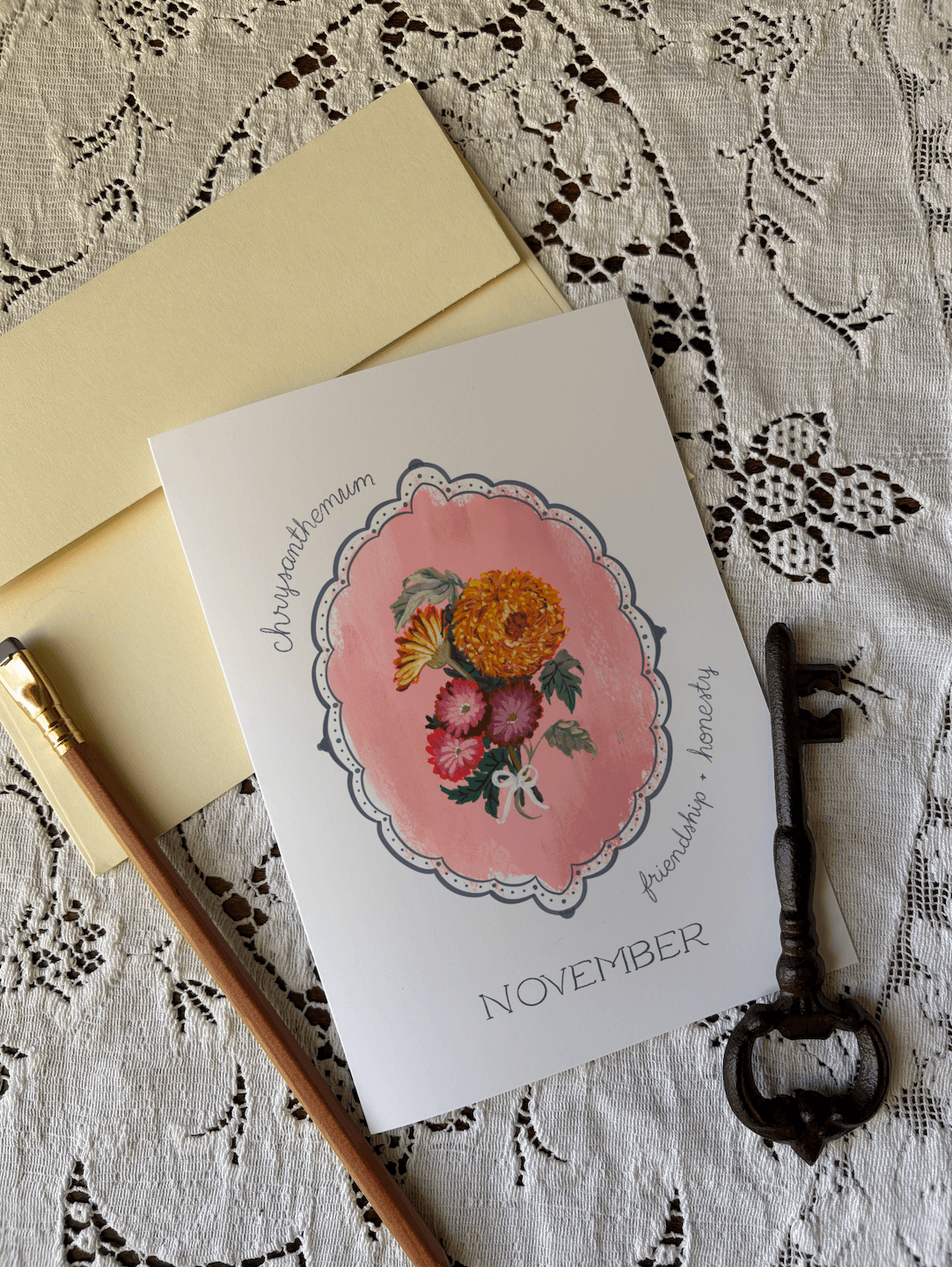 A flat-lay of a white greeting card featuring a pink floral medallion and the word 'NOVEMBER,' with a pale yellow envelope, a wooden pencil, and a vintage-style key on a white lace textile background.