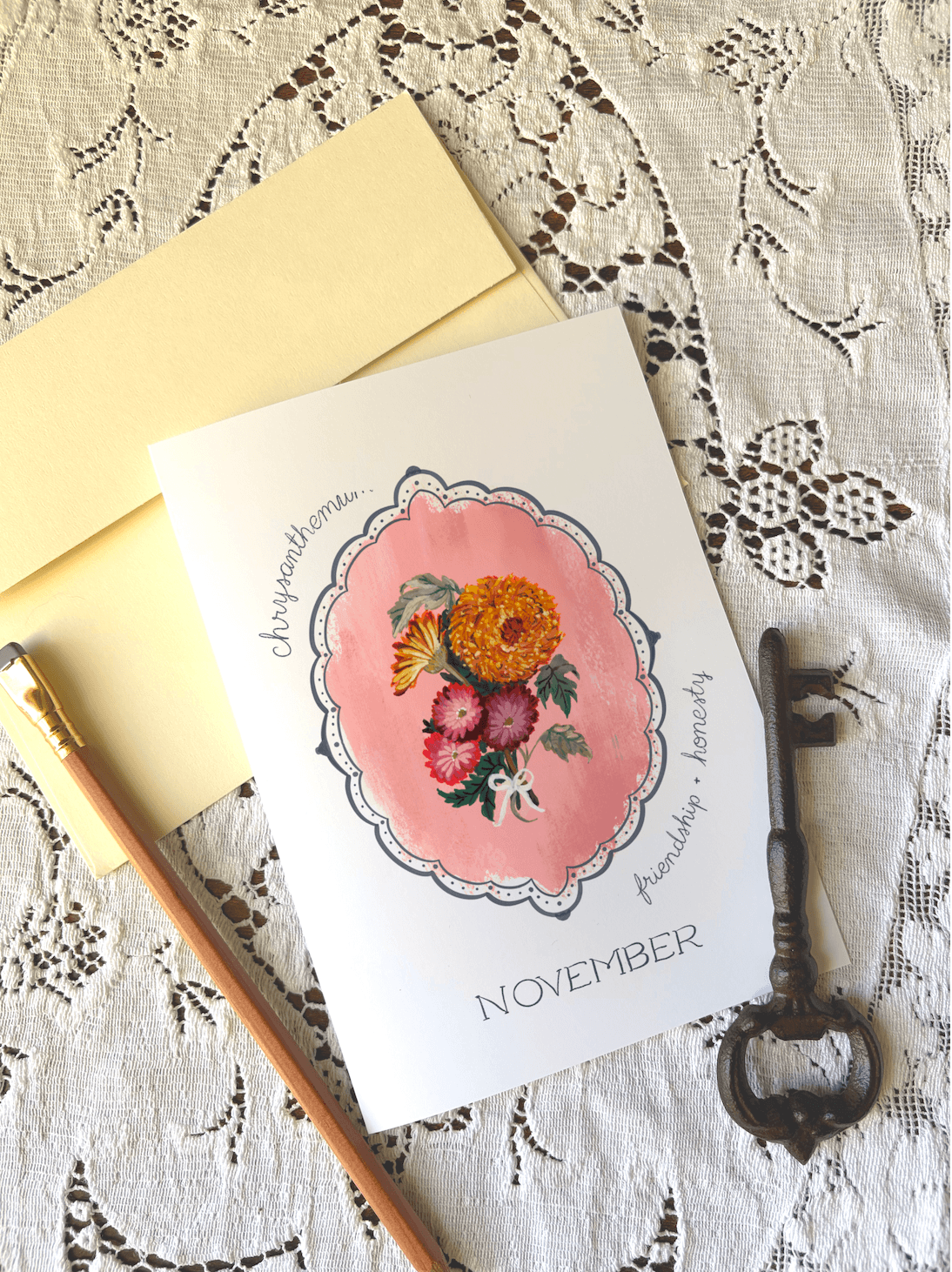 A flat-lay of a November card featuring a watercolor floral illustration in a pink frame, placed on a cream lace surface with a yellow envelope, a wooden pencil, and a vintage-style metal key.