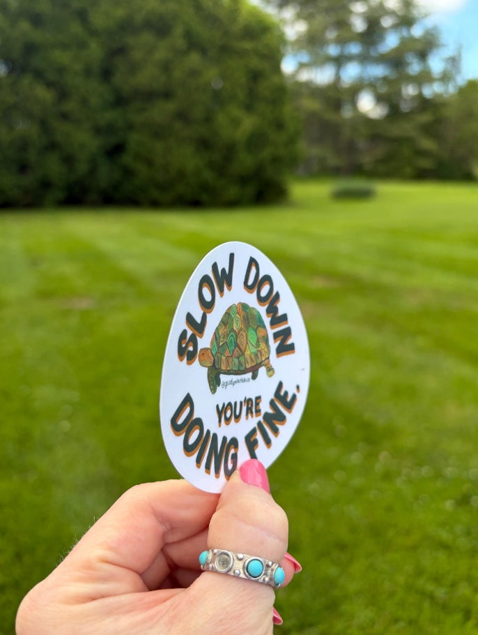 A hand with pink nail polish and a turquoise ring holds an oval white sticker that reads 'SLOW DOWN YOU'RE DOING FINE.' with a turtle illustration, against a blurred green lawn background.