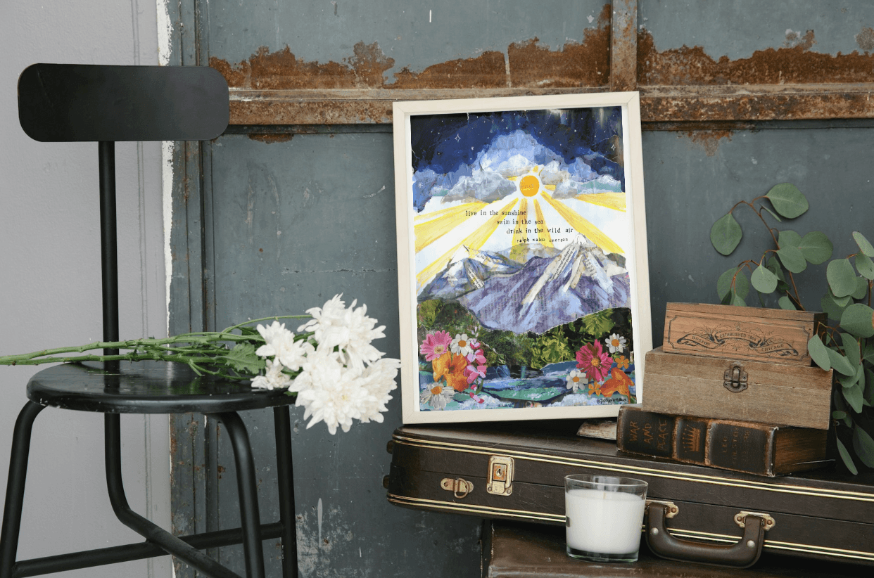 Artistic depiction of a mountain scene with flowers on a framed print, placed on a surface with a chair and books.