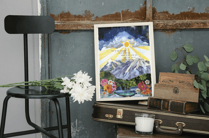 Artistic depiction of a mountain scene with flowers on a framed print, placed on a surface with a chair and books.