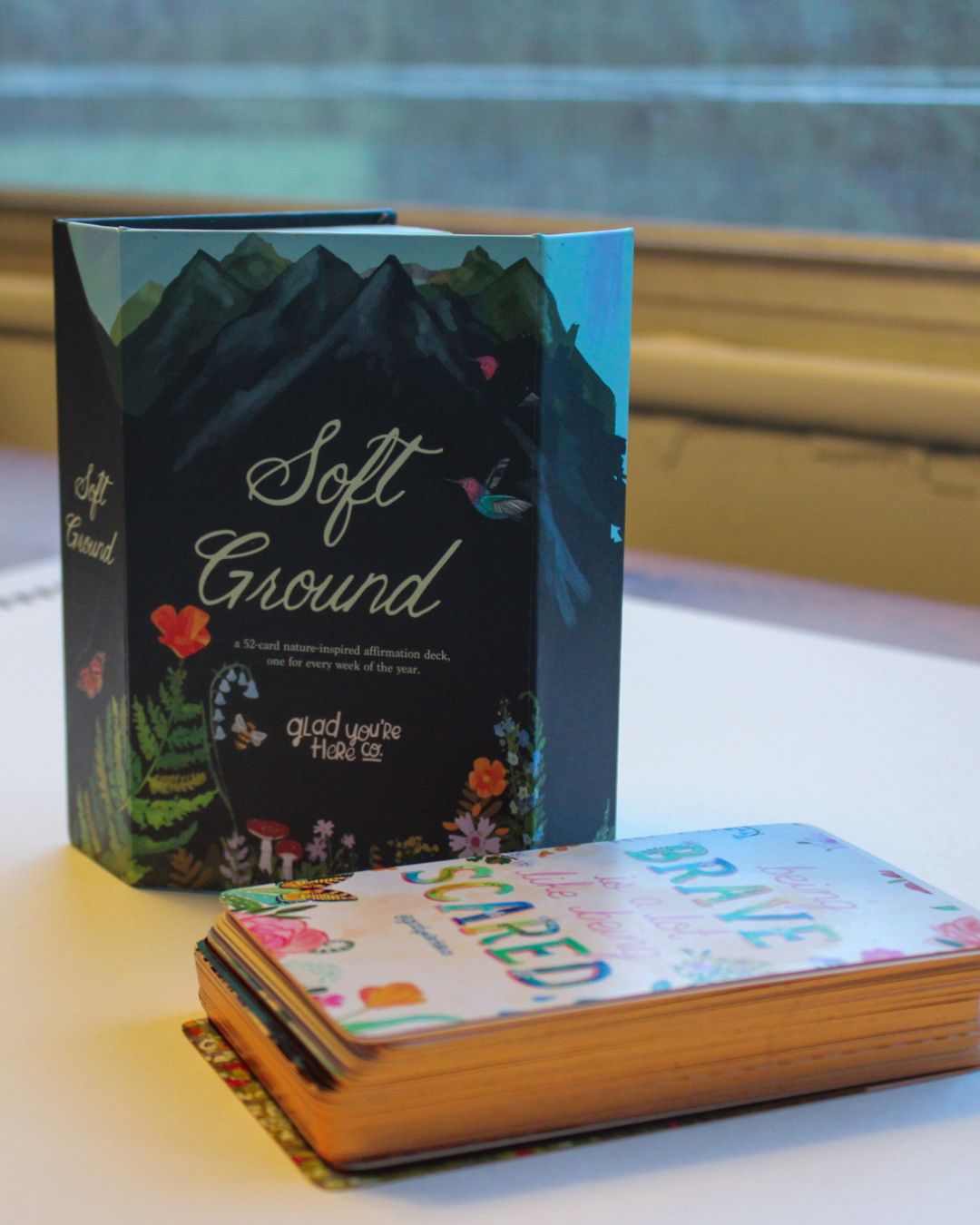 The 'Soft Ground' card deck box and a stack of cards on a tabletop near a window. The box features a dark mountain illustration with the words 'Soft Ground' and 'good you're Hard to see'.