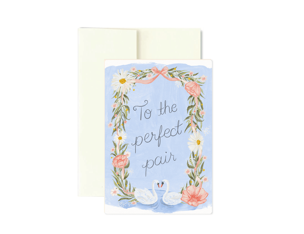 A square greeting card with a light blue background, featuring a floral wreath border, two swans, and the phrase 'To the perfect pair' in gray script. A cream envelope is placed behind the card on a white background.