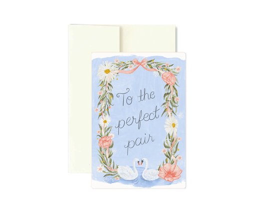 A square greeting card with a light blue background, featuring a floral wreath border, two swans, and the phrase 'To the perfect pair' in gray script. A cream envelope is placed behind the card on a white background.