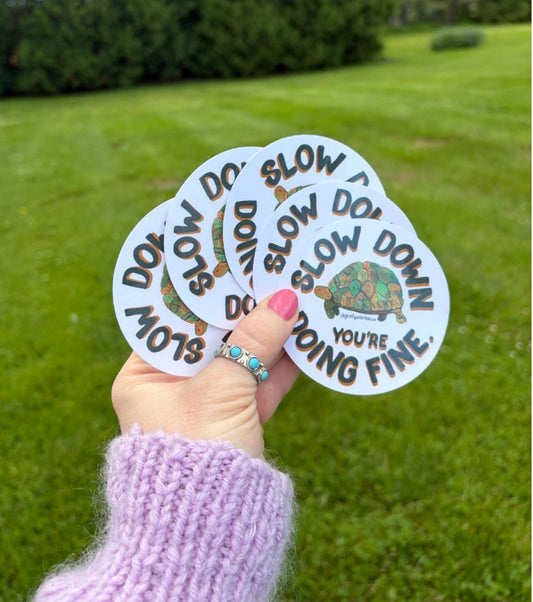 A hand wearing a lavender knit sweater holds a fan of five oval stickers outdoors on a green grassy lawn. The stickers feature the text 'SLOW DOWN YOU'RE DOING FINE' and  in brown lettering, with a small turtle illustration on one sticker.