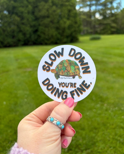 A hand with pink nail polish and a turquoise ring holds a round sticker in front of a blurred green lawn. The sticker features a turtle illustration and the text 'SLOW DOWN DOING FINE'.