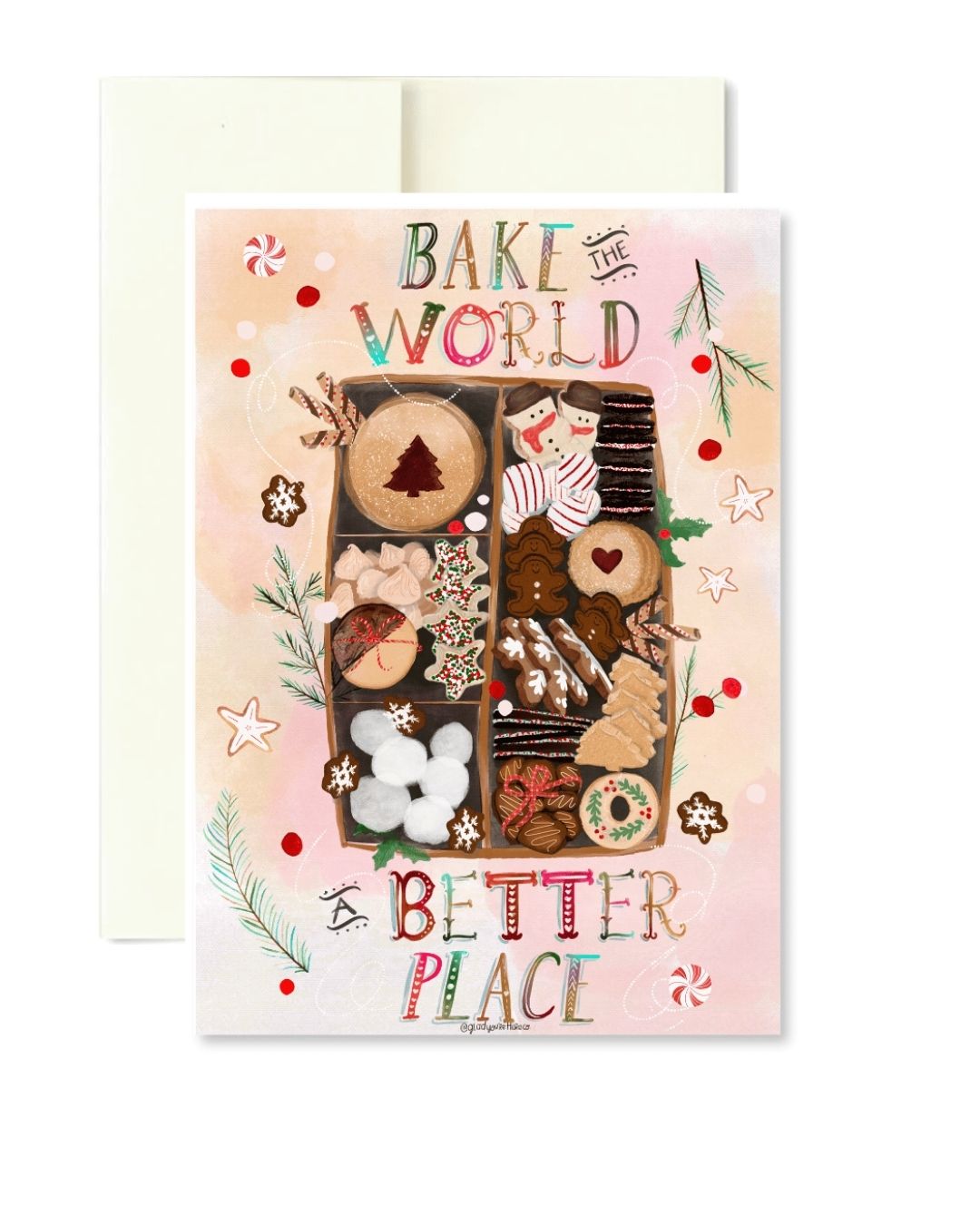A Christmas-themed greeting card with a pastel pink background, featuring an illustrated assortment of cookies and pastries arranged in a box, with the text 'BAKE THE WORLD BETTER PLACE' and a cream envelope behind it.