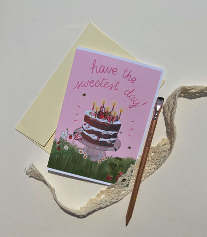 A pink greeting card with an illustration of a birthday cake and the words 'have the sweetest day!' in script, styled with a cream envelope, a wooden paintbrush, and a beige lace ribbon on a light background.