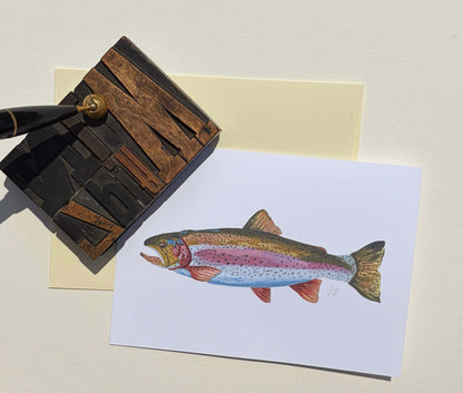 A white art card featuring a detailed watercolor illustration of a rainbow trout, styled on a cream background next to a wooden letterpress and a fountain pen.
