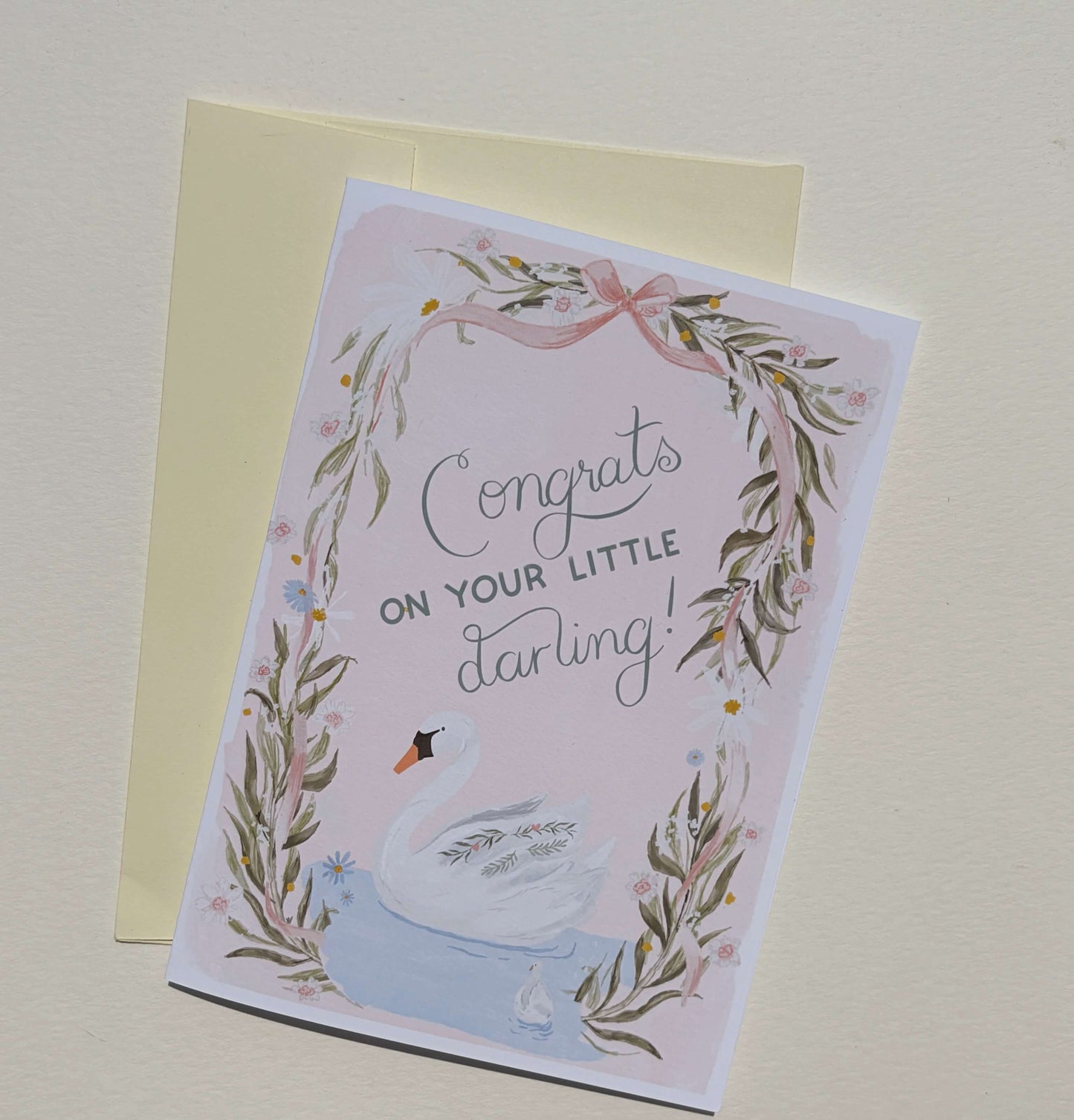 A congratulations card for a baby swan, featuring a watercolor illustration of a white swan in a blue pond, surrounded by a floral wreath and the text 'Congrats on your little darling!'. The card is shown with a pale yellow envelope on a light beige background.